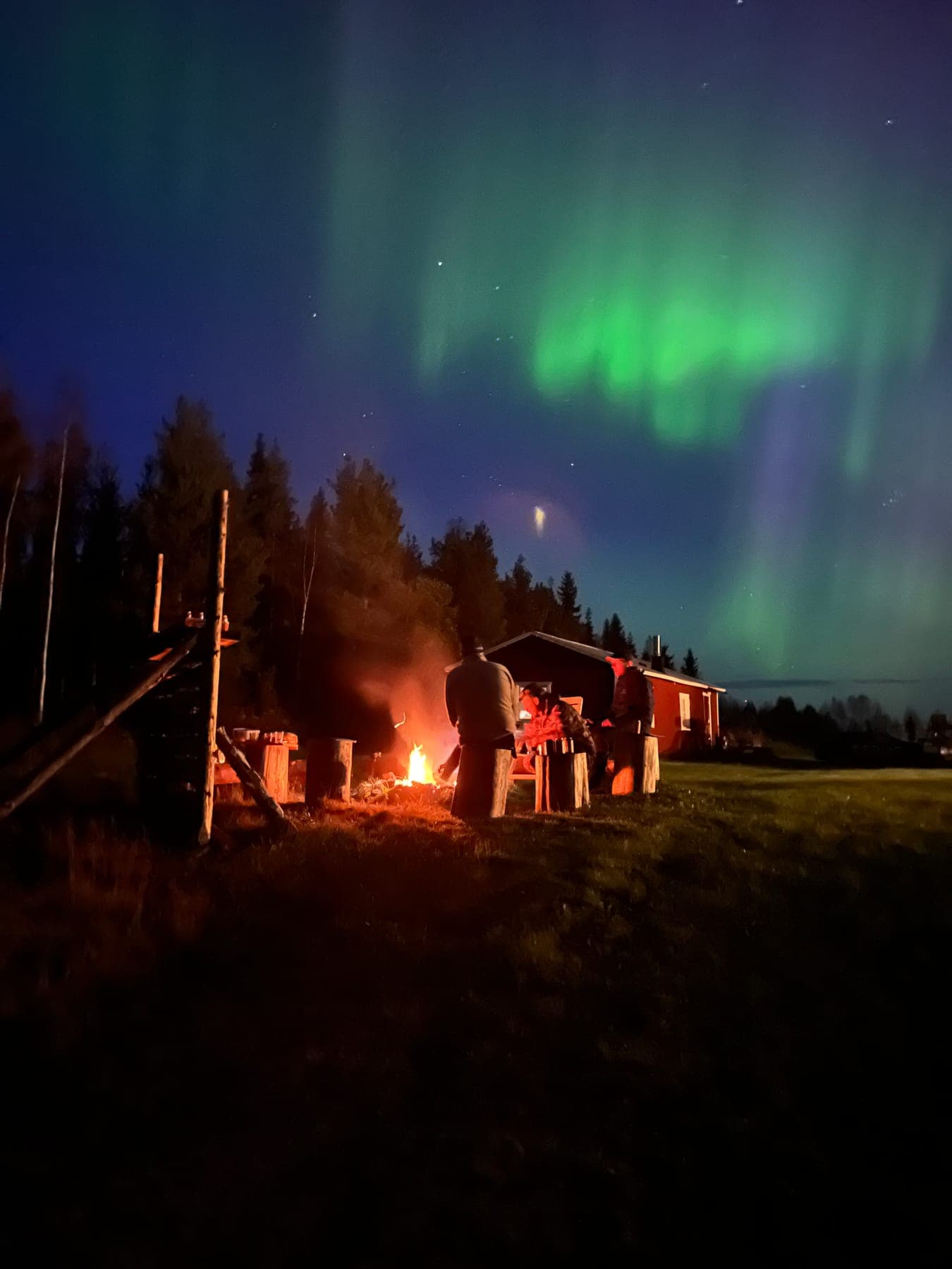 Sällskap kring lägereld under grönt norrsken